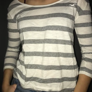 Randig tröja Hollister  - Vit 3/4-tröja med grå/glittriga ränder från Hollister, strl XS. Köpare står för frakt