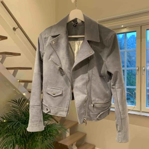  - Ljusgrå/blå (mer åt det grå hållet) jacka med mockaimitation. Är i jättefint skick, aldrig använd. Går att ha både inomhus och utomhus. Den är ganska kort, går inte över rumpan. Frakten är inkluderad i priset.