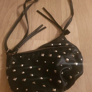  - Svart skinnimitation väska med nitar fram. Axelrems växa. Liten innerficka finns i andra facket.  Ena dragkedjan är sönder men går att laga. Mått ca. 26*17 cm Köparen betalar frakt. Samfraktar gärna med det andra jag säljer. 
