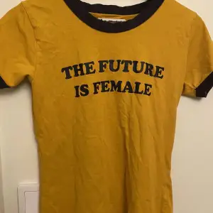 En gul tröja med svart runt armar och hals och tryck på tröjan där det står att framtiden är kvinnlig!