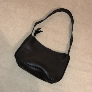 Svart väska - Väskans mått är cirka 22 cm bred och ungefär 12 cm hög. Materialet är lite glansigt.