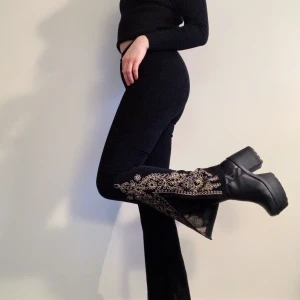 Faux Suede-byxor - Byxor med utsvängda ben i faux suede. Stängs med gylf och knapp. Höga i midjan. Utsmyckade med guldfärgade broderier och slits. Superfint skick. Midjemått 63 cm, längd 98 cm. Frakt 63kr.