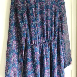 Blommönstrad, blå/lila klänning med öppen rygg, tunna ärmar och guldkedja som accessoar baktill. Från BIKBOK. Passar XS och S.