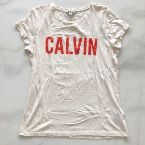 Calvin Klein Tshirt - Offwhite Calvin T-shirt med röd glansig text. Använd ca 2 gånger. Liten fläck vid nedre sömmen se bilder. Frakten är inräknad i priset!
