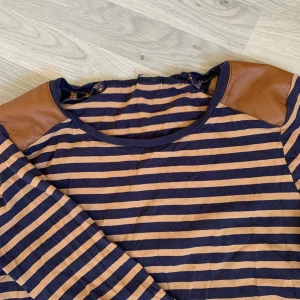 Långärmad, randig tröja - Blå och beige randig tröja med fake läder lappar på axlarna. Färgen är som på bild 1. Storlek står inte men skulle gissa på s/m. Köpare står för frakt. Kan mötas upp i Västerås/hallsta