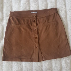 Kjol i mockaimitation - Superfin kjol i mockaimitation. Något stretchig i tyget. Säljes pga lite för stor för mig. Kanten i bak hänger ned litegrann (se sista bilden) men det går enkelt att fixa med tex ett stygn. Knappar hela vägen och fejk-fickor. Frakt ingår! :)