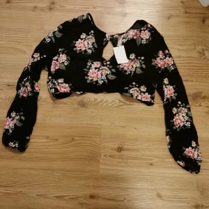 Svart blommig crop top. - En blommig långärmad crop top. Med två små knappar på baksidan, runt halsen och med knyt bak i ryggen. Helt ny aldrig använd, prislapp kvar
