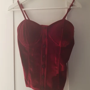  - Tröja i korsettstil, sammetimitation, mörkröd, aldrig använd. Liten i storleken men töjbar pga resor i ryggen. Vadderade kupor, mjuk och lätt att röra sig i. Justerbar remmar.