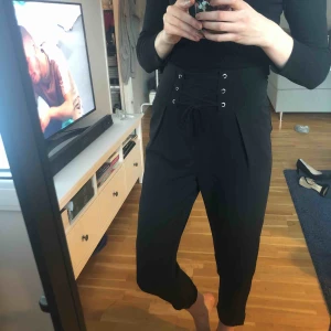  - Snygga byxor med fin snörning som detalj. Hög i midjan och lite kortare i benlängden. Knäpps med dragkedja i sidan. 