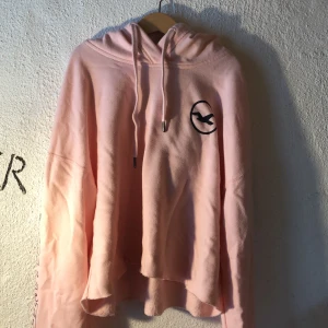 Hollister cropped hoodie - Rosa croppar hollister hoodie, tryck på huva och tryck på ärmarna.  Avklippt nederkant dok inte av migsjälv (köpare står för frakt)