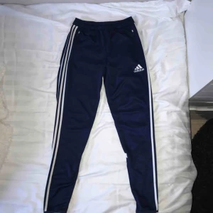  - Blå Adidas byxor med 3 sträck på sidan av benen och dragkedja vid benslut. 