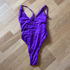 Lila baddräkt  - Lila baddräkt med djup rygg och high cut modell. Fina detaljer med rosetter på sidorna