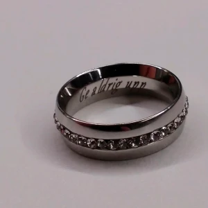 Stålring  - Obs! Graverad med "Ge aldrig upp". Helt oanvänd ring, tillverkad i 316L kirurgiskt stål med slipade cubic zirconia stenar runt hela. Ringen är i storlek 18,8 mm (passar dig som vill ha storlek 19). Den är kupad och 7 mm bred. Ringen är gjord i ett material som är väldigt hårt. Vilket gör att den behåller sin form trots smällar och påfrestningar. Köpare står för frakt 💜😋