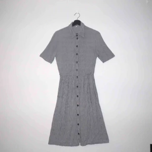  - Svart- och vitrutig klänning med knappar hela vägen och resår i midjan. Köpt på Beyond retro. Tunn och lätt. Gott skick.   Klänningen är 122 cm lång, 74 cm i midja (utan att resåren är uttöjd) och 46 cm i axelmått.