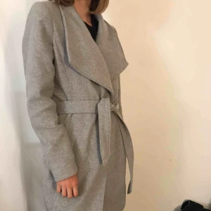  - Grå varm vinterjacka. Den är fodrad vilket gör den lämpligare för vintern. Den har ett skärp och knappar. Den är i gott skick. Den går ner till knäna ungefär. Nypris 999kr