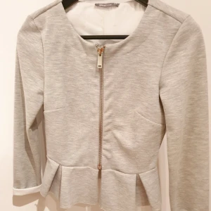  - Ljusgrå jack-kavaj med volang från Gina Tricot 