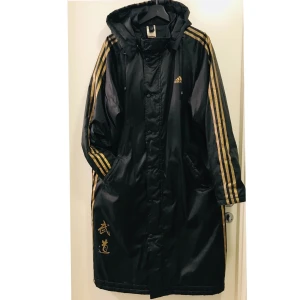 Adidas football coat  - Fotbollskappa i svart & guld. 115cm lång mätt från axeln. Baksidan utan tryck. Luvan är avtagbar. Lite slitet i den guldiga färgen på vissa ställen men annars i perfekt skick. Mellanvädersjacka. Skickas el hämtas i Malmö. 