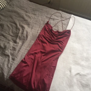  - Vinröd klänning i stretchigt, glansigt material från Nelly. Cowl-neck fram och djup urringning bak. Tunna axelband som korsas i ryggen. Aldrig använd, som ny.