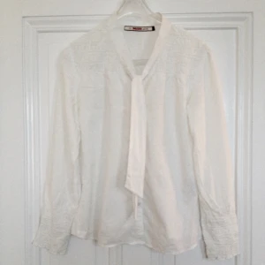  - Vit blus med knytsnörning i halsen, gör rosett eller ha som den är. Lätt puffärm och fina detaljer i ärmslut. Fint tunt material med insydda prickar. Nyskick.  FRI FRAKT!