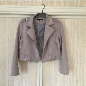 Lila/grå jacka i mocka st 38/40 - En pastell lila/grå midjejacka i mockatyg. Använd bara en gång så i nyskick. Silvriga dragkedjor för fickor framtill. Passar en st 38/40. Mått: armhåla-armhåla: 45 cm. Längd från axel till midja (uppifrån och ner): 42 cm. 