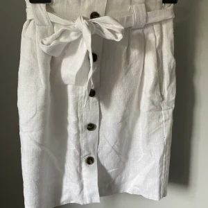 Vit kjol med knytband  - En vit kjol i tunt material. Kjolen är längre i modellen. Den har ett  knytband i midjan som man kan dra åt så att den kan passa många. Finns även en liten vit dragkedja på sidan och har två stora fickor på varsin sida. Helt oanvänd och köptes för 250kr men säljs nu för 115kr.