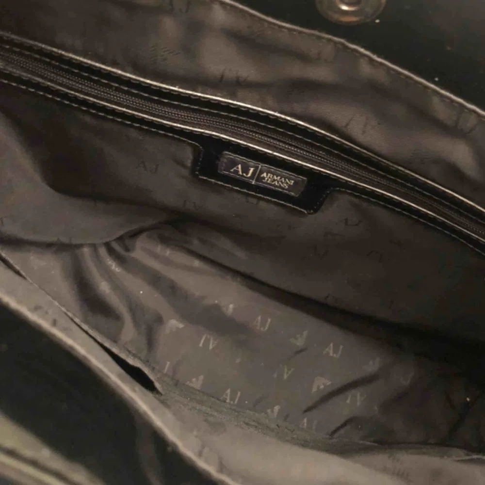 Handväska från Armani Jeans har en svart lackad yttre präglad med Armani jeans logotyp och en matchande metallbricka. En magnetlås visar en textil inre med flera fack och en dragkedja I fint begagnat skick Nypris 1200:- Mitt pris 600:-. Väskor.