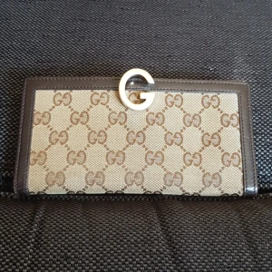 Plånbok från Gucci - Skinnplånbok med gucci märket i tygdetaljer på utsidan. Flertal fack inuti samt ett myntfack på baksidan. Låses med ett guldfärgat metal G som väldigt snygg detalj. 