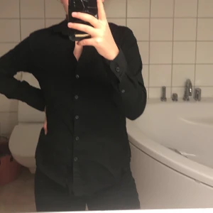 Svart skjorta - En väldigt basic svart skjorta. Säljer pga att den är för stor över brösten på mig (85B). Den är ganska öppen upptill. Går att styla som man vill då den har 5 knappar på magen och 3 knappar på vardera mudd på ärmarna. Skriv för fler bilder. Jag är ca 170cm lång och väger 70kg. Betalning sker endast via Swish!