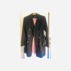 Svart Klassisk Trenchcoat Stl 36 - Svart Trenchcoat med snygga detaljer vid axlar/ok. Ficklock. Minimalt slitage. Använd max 5 ggr. Längd nacke/fåll 85 cm. Köparen betalar frakten.
