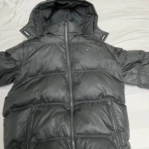 Tommy Hilfiger Svart Essential Down Jacket - Svart jacka från Tommy Hilfiger. I använd skick. Jackan har en avtagbar luva och stängs med dragkedja.Bekväma fickor.Köpt från Kidsbrandstore. Storlek: 164 cm (14 år).Produktbeskrivning. Jacka, Luva, Fickor, Resår, Brodyr, Patch, Material: 100% Polyester.