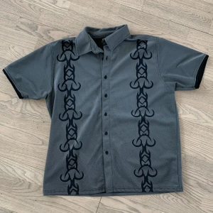 Grå 90-tals skjorta!  - En grå kortärmad skjorta i polyestertyg, relativt luftig. Har ett snyggt mönster på framsidan. Det står XL, men sitter mer som en liten L/stor M. Säljer då den ej passar min stil längre!  Plagget har inga skador. Kan mötas upp i Gbg!