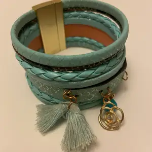 Ett turkost armband i läderimitation. Armbandet består av sju delar i olika mönster och flätor med silverdetaljer. Det har öglor med hängdetaljer som en tofs i tyg, en blå pärla och en ros i guldfärg. Armbandet stängs med hjälp av en magnet Begagnat skick