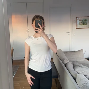 Vit T-shirt låg rygg - Vit dam T-shirt med tightpassform figursydd. Tunt tyg i 95% bomull. Fri från fläckar och missfärgning. Lågrygg har i dessa bilder en sport-bh under plagget som syns i ryggen.