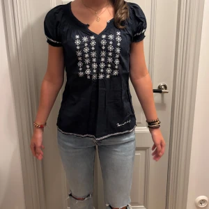 Mörkblå blus med broderi från Bondelid - Supersöt mörkblå blus från Bondelid med vita broderade detaljer framtill, på ryggen och på ärmarna. Blusen har korta puffärmar, rung halsringning med en liten knapp och ett unikt mönster både fram och bak 🥰