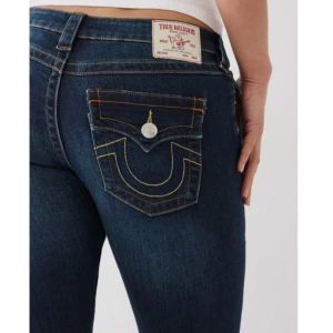 True Religion mörkblå bootcut jeans - Snygga Joey low rise flare jeans från True Religion med bootcut passform och låg midja. Jeansen har ikoniska bakfickor med gul kontrastsöm och silverfärgad knapp. Materialet är klassisk jeans med stretch för extra komfort. Perfekt för dig som gillar en cool och tidlös look.