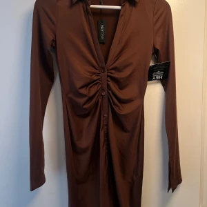 Brun långärmad klänning från NLY One - Snygg brun långklänning från NLY One med lång ärm och krage. Klänningen har en cool rynkad detalj framtill med knappar längs mitten och slits vid ärmslut. Materialet är mjukt och stretchigt, perfekt för dig som gillar en slim passform och stilren look.
