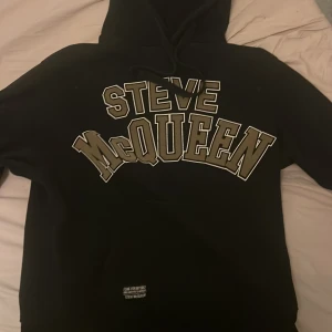 Svart Steve McQueen hoodie - Svart hoodie från Steve McQueen med stor, kontrasterande logotyp i vitt och beige på bröstet. Tröjan har huva med snörning och klassisk känguruficka framtill. Perfekt för dig som gillar streetwear och vill sticka ut med en snygg logga.