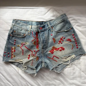 Levi's jeansshorts med studs - Unika ljusblå jeansshorts från Levi's med coolt broderi samt nitar på fram- och bakfickor. Rå kant nedtill och slitna detaljer ger en edgy vibe. Klassisk femficksmodell med knappgylf och midwaist passform. Aldrig använda. W28.