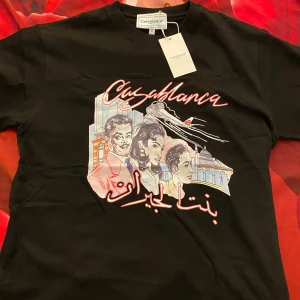 Svart Casablanca t-shirt med tryck - svart t-shirt från Casablanca med färgglatt retroinspirerat tryck framtill som visar tecknade figurer, byggnader och flygplan. T-shirten har rund hals och korta ärmar. Perfekt för dig som vill sticka ut med en unik design.