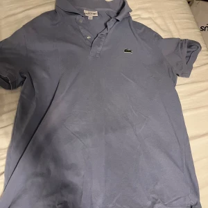 Ljusblå Lacoste slim fit piké - Snygg ljusblå pikétröja från Lacoste i slim fit-modell. Klassisk krage, två knappar och den ikoniska gröna krokodilloggan på bröstet. Tillverkad i bomull med korta ärmar, perfekt för en chill och stilren look.