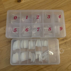 nageltippar i olika storlekar - Två askar med nageltippar i olika storlekar, både transparenta och vita. Nageltipparna är gjorda av plast och kommer i organiserade fack med siffror för enkel användning. Perfekt för att skapa egna naglar hemma eller till nagelkonst. INTE HELT NYA dvs inte fullt med tippar men finns tillräckligt till flera set
