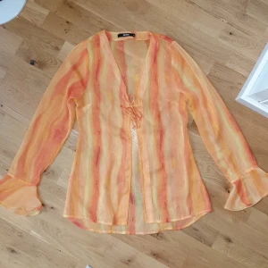 Meshtopp / blus i orange toner - Genomsnittlig meshtopp / sheer blus från Bikbok i storlek XS med v-ringning och snörning framtill. Blusen har långa ärmar med volang vid ärmslut och ett randigt mönster i orange, gul och röda nyanser. ⭐ Använd 1 gång, passar storlek XS-S.   📷  Inspiration: https://www.zalando.se/lipsy-strandaccessoar-pink-print-li781t01m-j11.html