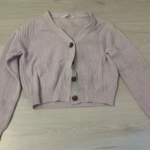 Ljuslila kofta från H&M med knappar - Säljer en ljuslila, ribbstickad kofta från H&M med stora mörka knappar framtill. Koftan har v-ringad hals, croppad passform och långa ärmar. Perfekt att slänga över en t-shirt för en chill look. Materialet är mjukt och stickat.