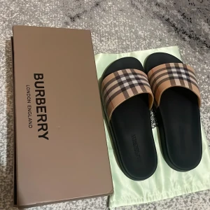 Burberry sandaler med rutig mönster - Snygga sandaler från Burberry med klassiskt rutig mönster i beige, svart och rött på remmen. Sulan är svart och har en platt form med Burberry-logga präglad på fotbädden. Perfekta för sommaren och ger en lyxig vibe till din outfit.