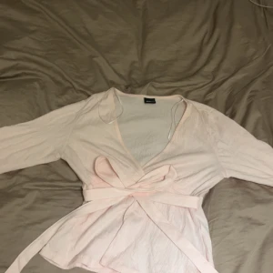 Ljusrosa omlottopp med knytband - Säljer en ljusrosa omlottopp från Gina trikot med långa ärmar och v-ringad hals. Toppen har ett brett knytband i midjan som ger en snygg siluett. Perfekt för dig som gillar mjuka färger och stilrena detaljer.