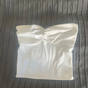 Vit bandeau tubtopp från Bik Bok - Snygg vit bandeau tubtopp från Bik Bok i storlek S. Toppen har en twistad detalj framtill och smockad baksida för extra komfort. Materialet är mjukt och har en lätt glansig finish som ger en fräsch look. Perfekt att styla med jeans eller kjol för en trendig vibe.