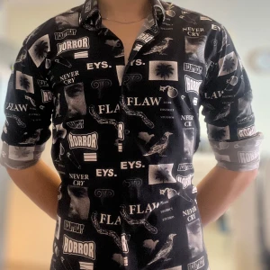 Svart mönstrad skjorta Moschino - Unik svart skjorta från Moschino med coolt tryck i vitt och grått med text, palmer, kedjor och fåglar. Skjortan har klassisk krage, långa ärmar och knappar framtill. Perfekt statement-plagg för dig som gillar edgy stil och grafiska prints.