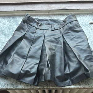 skinnkjol från Zara - Cool svart plisserad kjol från Zara i skinnimitation med bred linning och hällor för bälte. Kjolen har en kort och utsvängd modell som ger en edgy vibe. Perfekt för att lyfta din outfit med lite attitude.