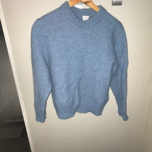 Blå stickad tröja med rund hals - Mysig blå stickad tröja med rund hals och långa ärmar. Tröjan har en enkel design utan mönster och är tillverkad i ett fluffigt material som känns skönt mot huden. Perfekt för att hålla värmen under kyliga dagar. Tröjan har slits på baksidan och vid slutet på armarna. Inte använd mycket och inget fel heller på den❤️