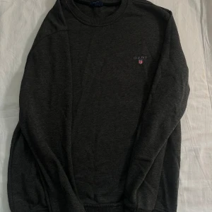 Mörkgrå sweatshirt från GANT - Säljer en stilren mörkgrå sweatshirt från GANT med broderad logga och emblem på bröstet. Tröjan har rund hals, ribbade muddar och lång ärm. Perfekt för dig som gillar klassisk och enkel design med en touch av preppy stil.